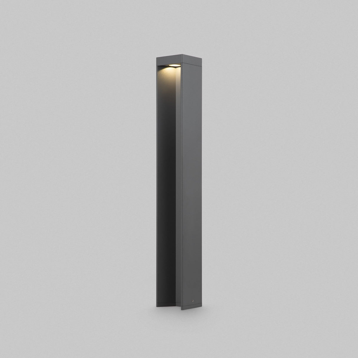 The Essen LED Bollard Light 65cm in dark gray, with its tall and sleek design, stands against a light gray backdrop. It features a corrosion-resistant square base and emits a soft glow downward from the small light at the top.