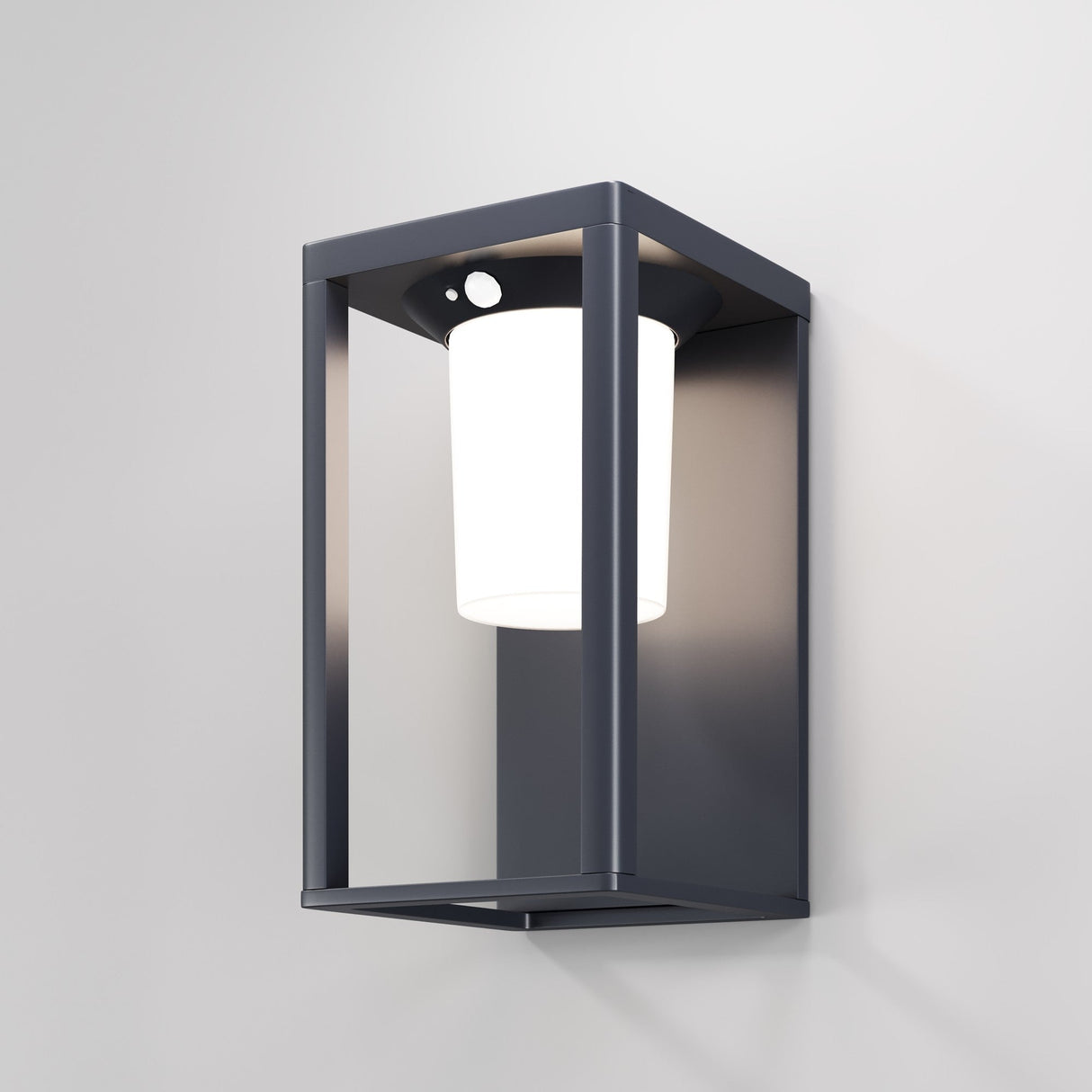 The Shatter Outdoor Wall Light 3000K - Graphite boasts a modern geometric design with a graphite metal frame and cylindrical white diffuser. Equipped with a solar battery, it attaches to a light-colored wall, casting a soft glow.