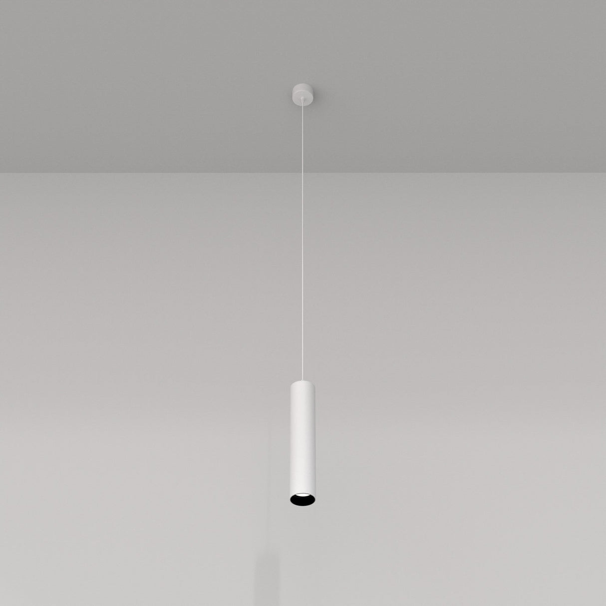 The Focus Single Pendant Light - White features a sleek, minimalist design that gracefully hangs from the ceiling, casting subtle shadows on the wall. Ideal for modern interiors, its cylindrical shape adds elegant charm to a plain gray backdrop.