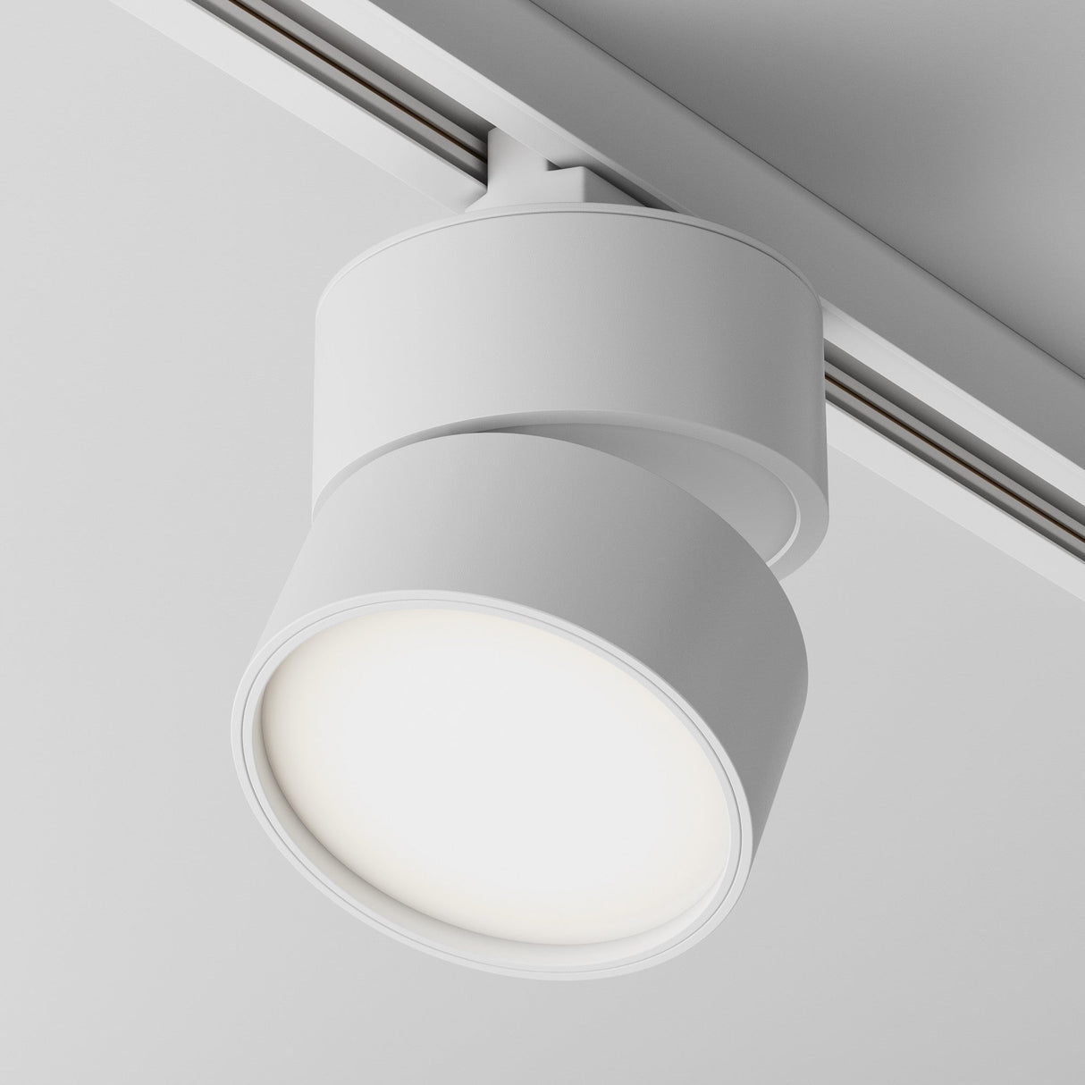 A close-up of the Onda LED Single Phase Unity Track Spotlight 12W 4000K - White shows its cylindrical design mounted on a metal track, emitting neutral white light with a 120° beam angle, casting a warm ambiance against a plain white ceiling.