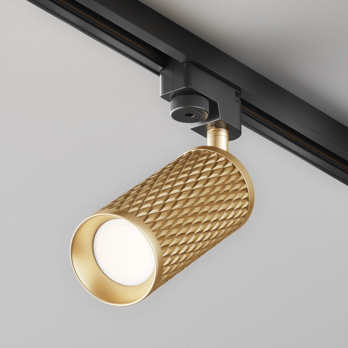 The Focus Design Single Phase Unity Track Spotlight in gold features a textured diamond pattern, mounted on a sleek black rail of the Unity track system against a white ceiling, directed downward to showcase its elegant design.