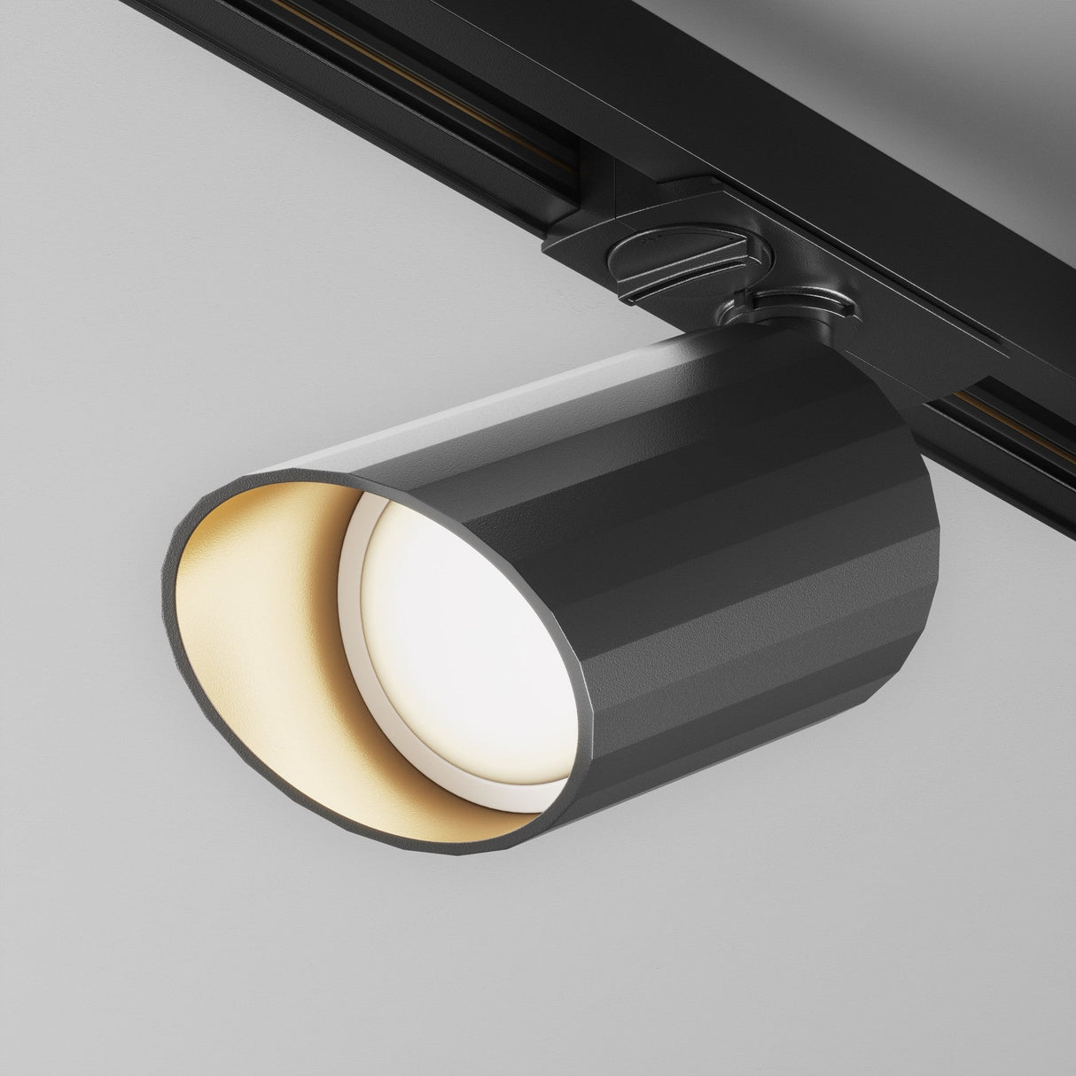 A close-up of the Lipari Single Phase Unity Track Spotlight in black, fixed to a ceiling. The light is on, emitting a soft, warm glow with a white inner surface. Its adjustable head enhances the sleek, minimalist design.