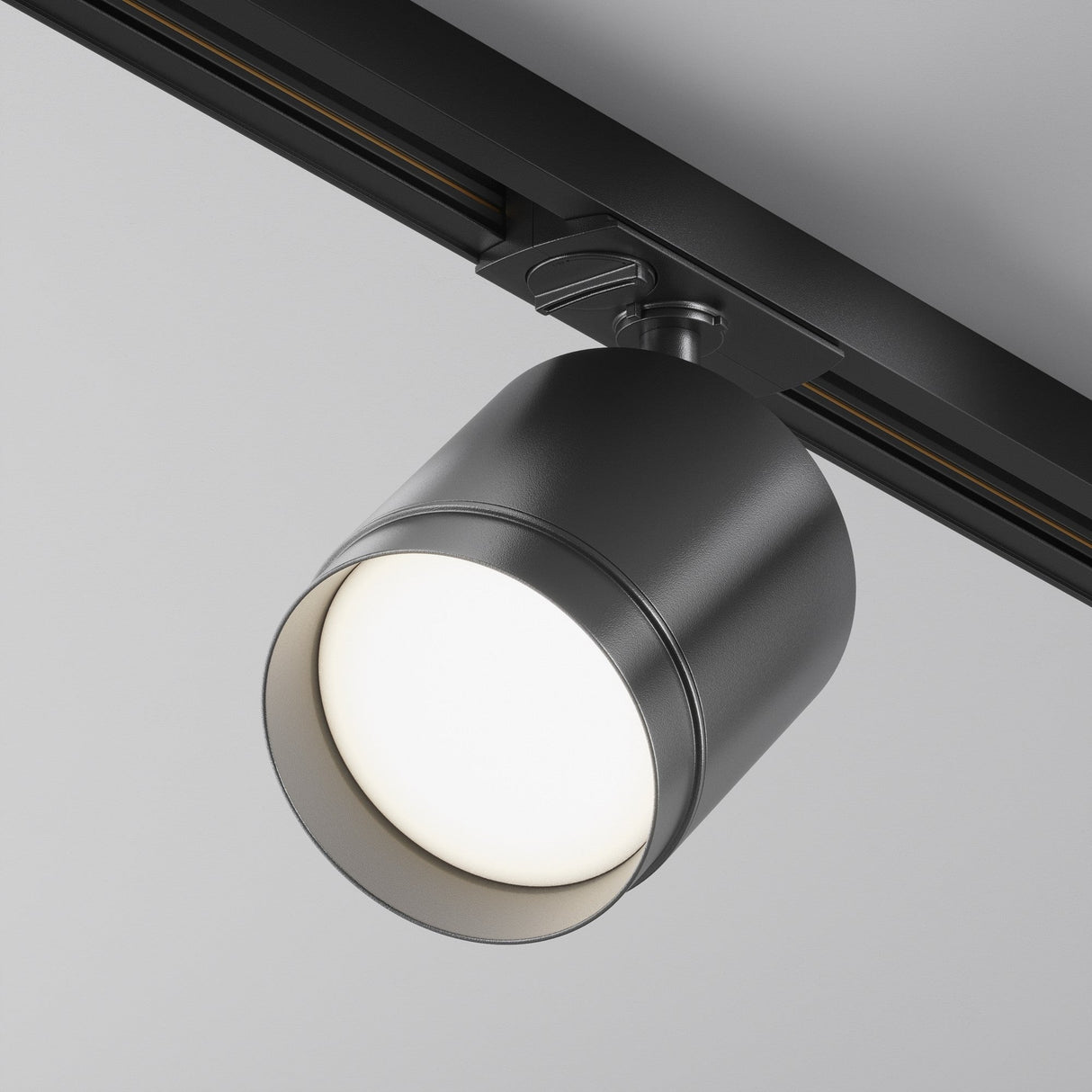 The Polar LED Single Phase Unity Track Spotlight - Black features a modern cylindrical design with a silver metallic finish. Mounted on a black track against a white background, it emits a bright white glow, providing elegant accent lighting for any space.