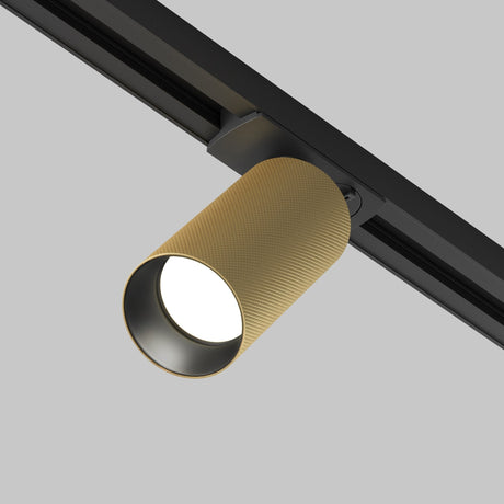 The Artisan Single Phase Unity Track Spotlight, featuring a cylindrical matte gold finish with ribbed texture, is mounted on a sleek black track against a light gray background, emitting a bright glow.