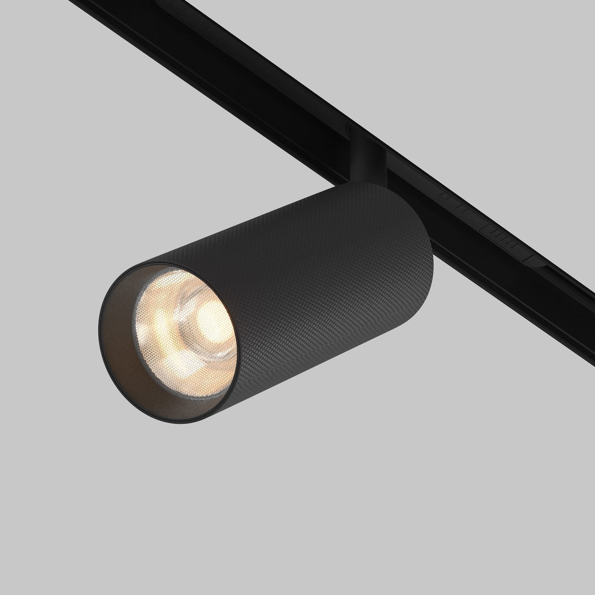The Artisan LED Magnetic Exility Track Spotlight 12W 3000K in sleek black, with a cylindrical design and anti-glare filter, is mounted on a ceiling rail against a gray background, casting a warm glow that enhances its modern minimalist elegance.