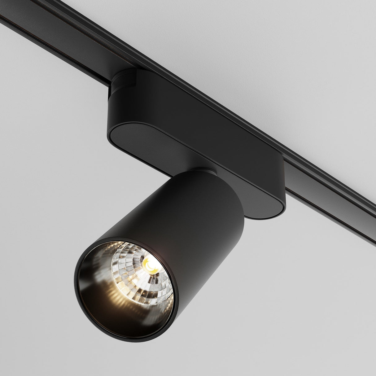 A close-up of the Focus LED Magnetic Radity Track Spotlight 12W 3000K - Black on a white ceiling, showcasing its cylindrical design and glowing bulb aimed downward. Ideal for accent lighting in modern interiors, it merges functionality with style.