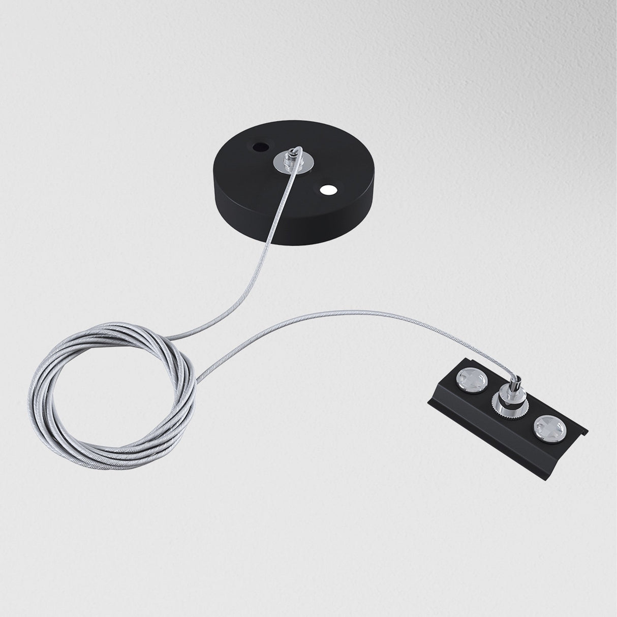 The Unity Single Phase Surface-Mounted Track Suspension Cable - Black, with its black circular base and rectangular component connected by a coiled suspension cable, evokes modern elegance against a plain white background.