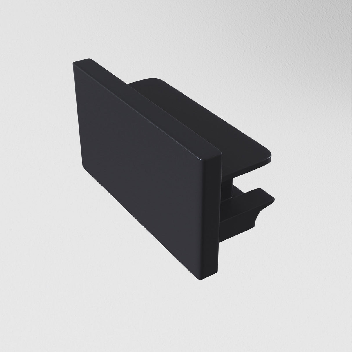 A black Unity Single Phase Surface-Mounted Track End Cap is beneath a minimalist, rectangular wall hook on a white wall. The hook features a flat, sleek surface and compact design.