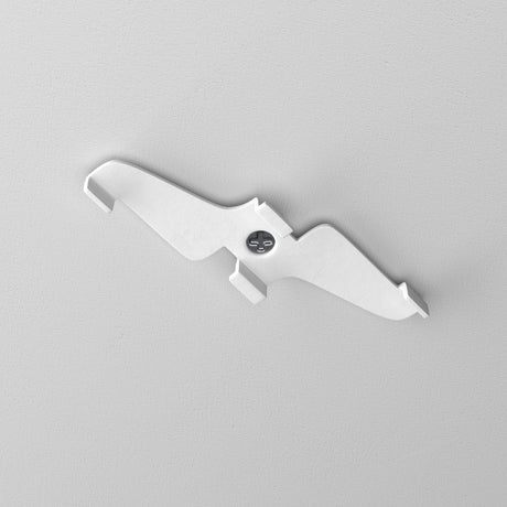 The Exility Magnetic Track Clamp - White creates a striking silhouette against the gray wall. Its minimalistic, bird-shaped design with outstretched wings harmonizes seamlessly with modern lighting, secured by a central screw for an elegant touch.