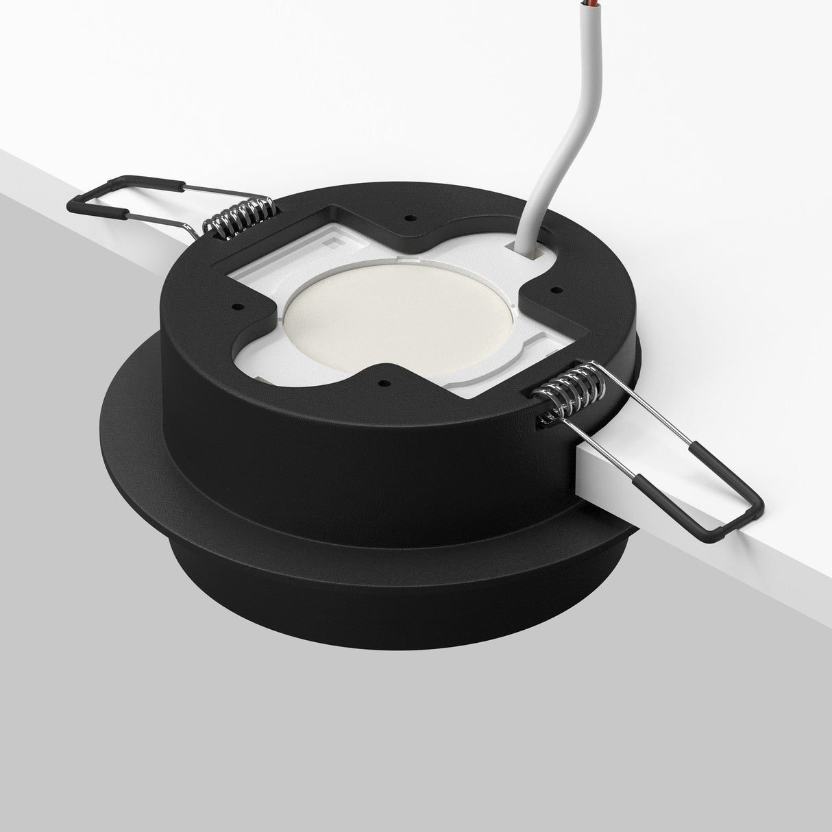 A close-up view shows an Orlo Fixed Round Recessed Downlight in matte black partially inserted into a white ceiling, featuring two metal spring clips on the sides and a white power cable extending from the top, perfect for an industrial decor vibe.