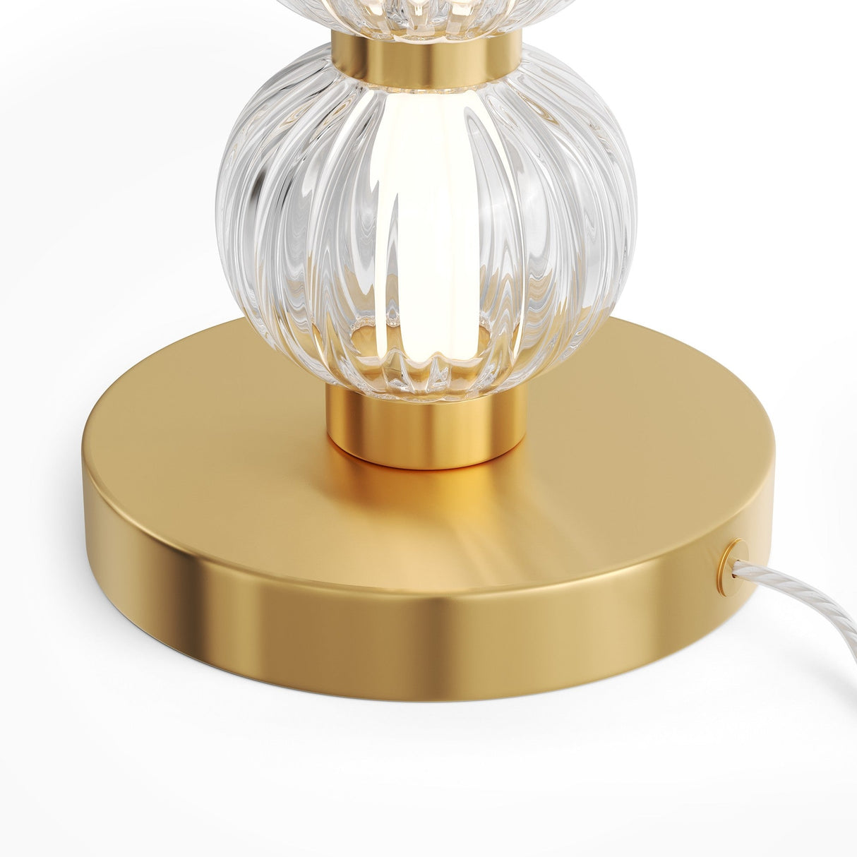 A close-up of the Amulet LED Table Lamp in gold shows ribbed glass spheres on a round base, with a white power cord on the side. The lamp emits a soft glow through its elegant glass design.