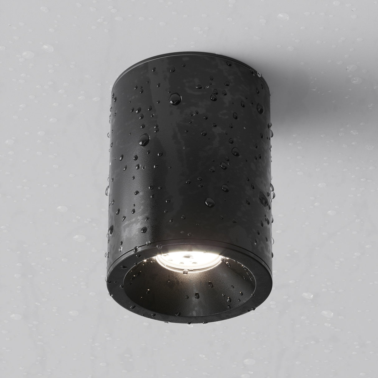 The Zoom Round Surface Downlight - Black, an IP65 cylindrical outdoor fixture, is wall-mounted and adorned with water droplets as it casts a downward glow. The light gray background enhances the elegant simplicity of this durable installation.