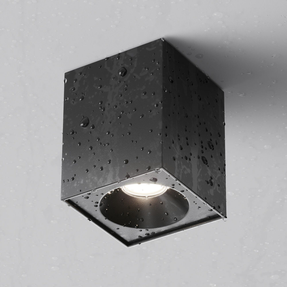 The Zoom Square Surface Downlight - Black, boasting an IP65 protection rating and matte black finish, elegantly mounts on a gray wall with water droplets adorning its surface as it shines downward.