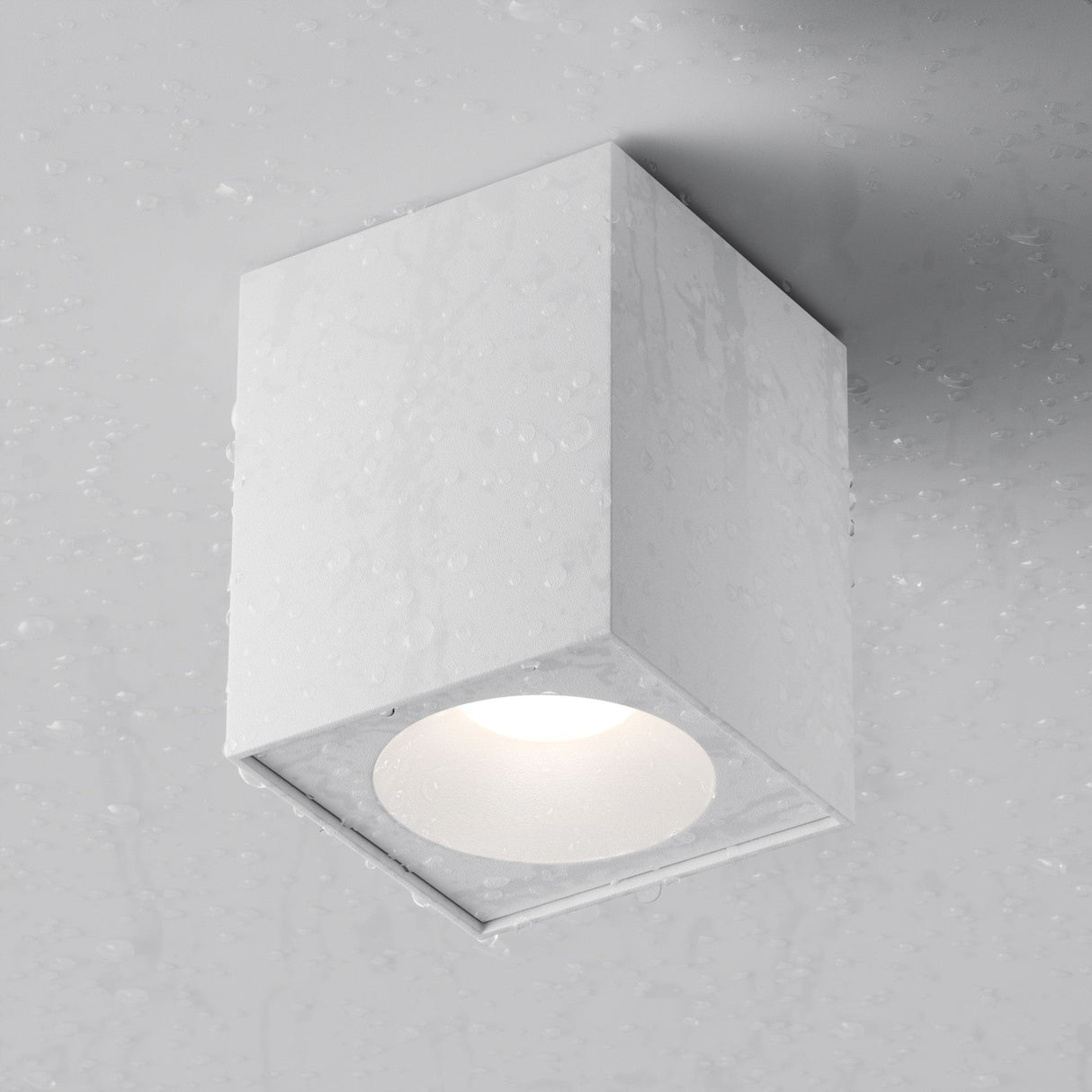The Zoom Square Surface Downlight - White is a minimalist ceiling fixture with an IP65 rating, ideal for modern interiors. Its square design attaches to a light gray ceiling, emphasizing its suitability for damp conditions with illumination and water droplets.