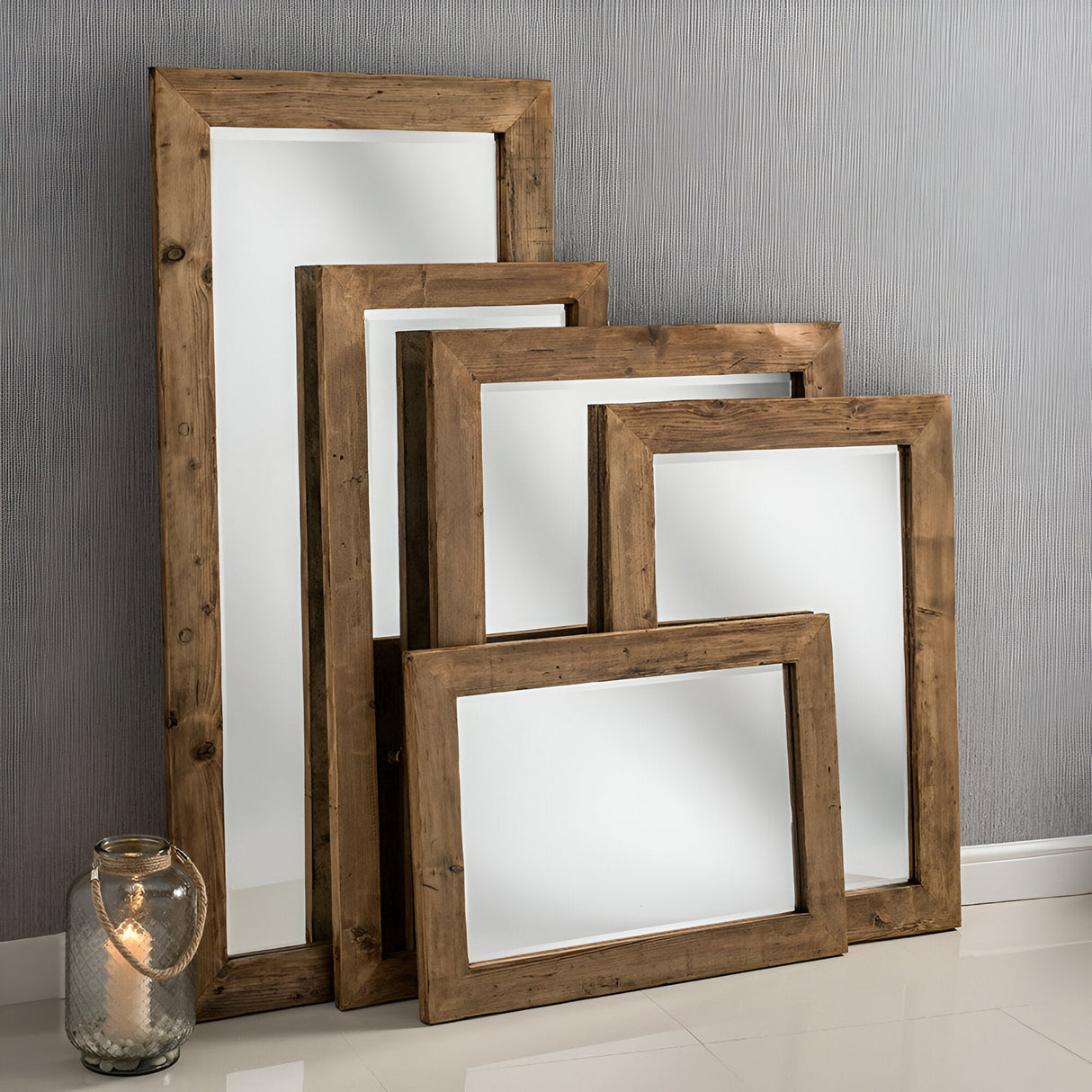 Four Glimpse Rectangle Wall Mirrors - Oak of different sizes are leaned against a light gray, textured wall. Embracing modern farmhouse style, a glass jar candle holder with a lit candle is placed on the white floor next to the mirrors, adding charm to this rustic home decor.
