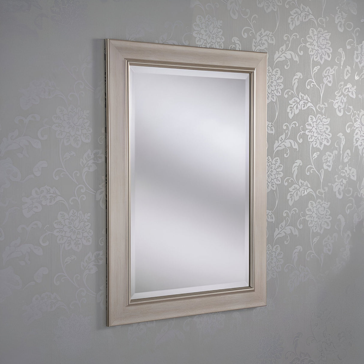 The Rimini Rectangle Wall Mirror - Silver, featuring a light wooden frame, is mounted on a wall adorned with floral-patterned wallpaper, enhancing the contemporary decor with its reflective surface.