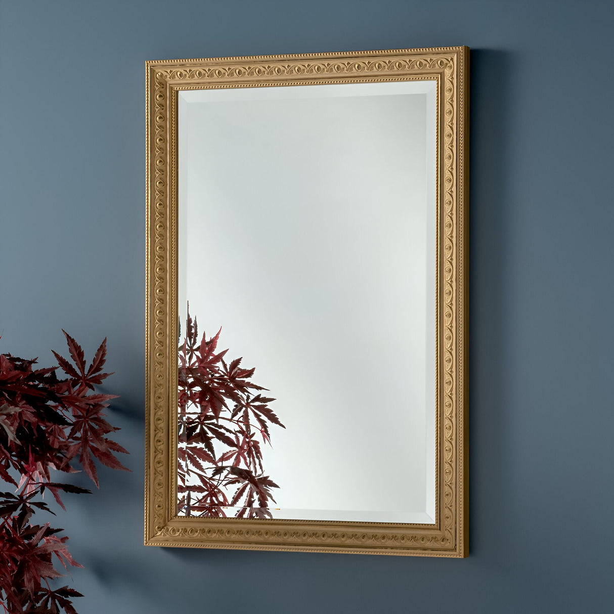 A Rocco Rectangle Wall Mirror - Gold is mounted on a blue wall, exemplifying luxury decor. The reflection captures maroon-colored leaves from a plant, bringing nature into the scene. This mirror's elegant design features intricate decorative details.
