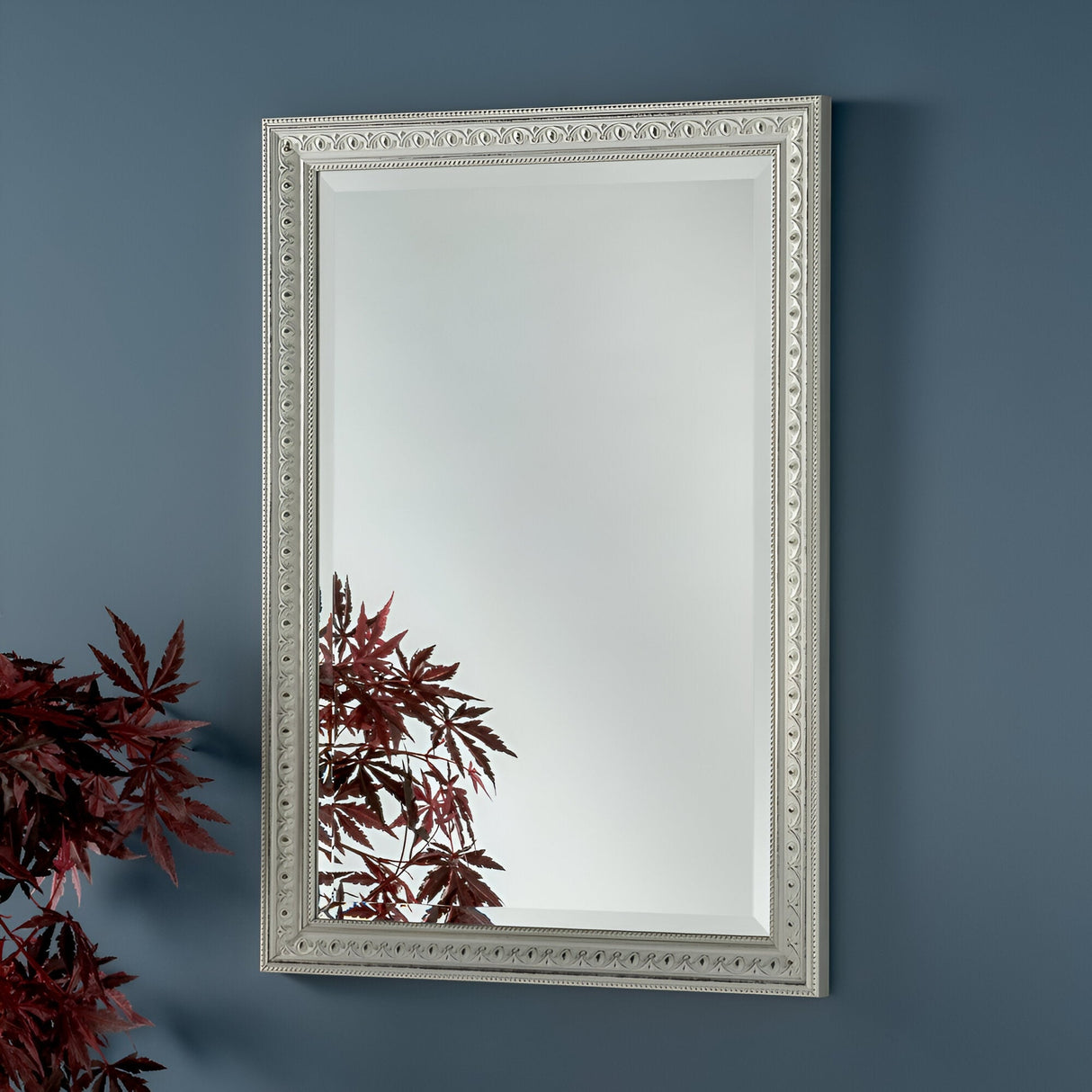 A Rocco Rectangle Wall Mirror with an ornate silver frame hangs on a blue wall. The reflection reveals red and burgundy foliage in the lower left corner, adding contrast to the minimalistic setting of this modern interior.