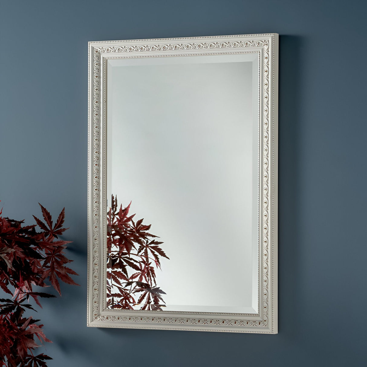 The Rocco Rectangle Wall Mirror - White is stylishly placed on a blue-gray wall, featuring its intricate frame. The reflection beautifully showcases the top of a decorative plant with dark red leaves, epitomizing modern simplicity.