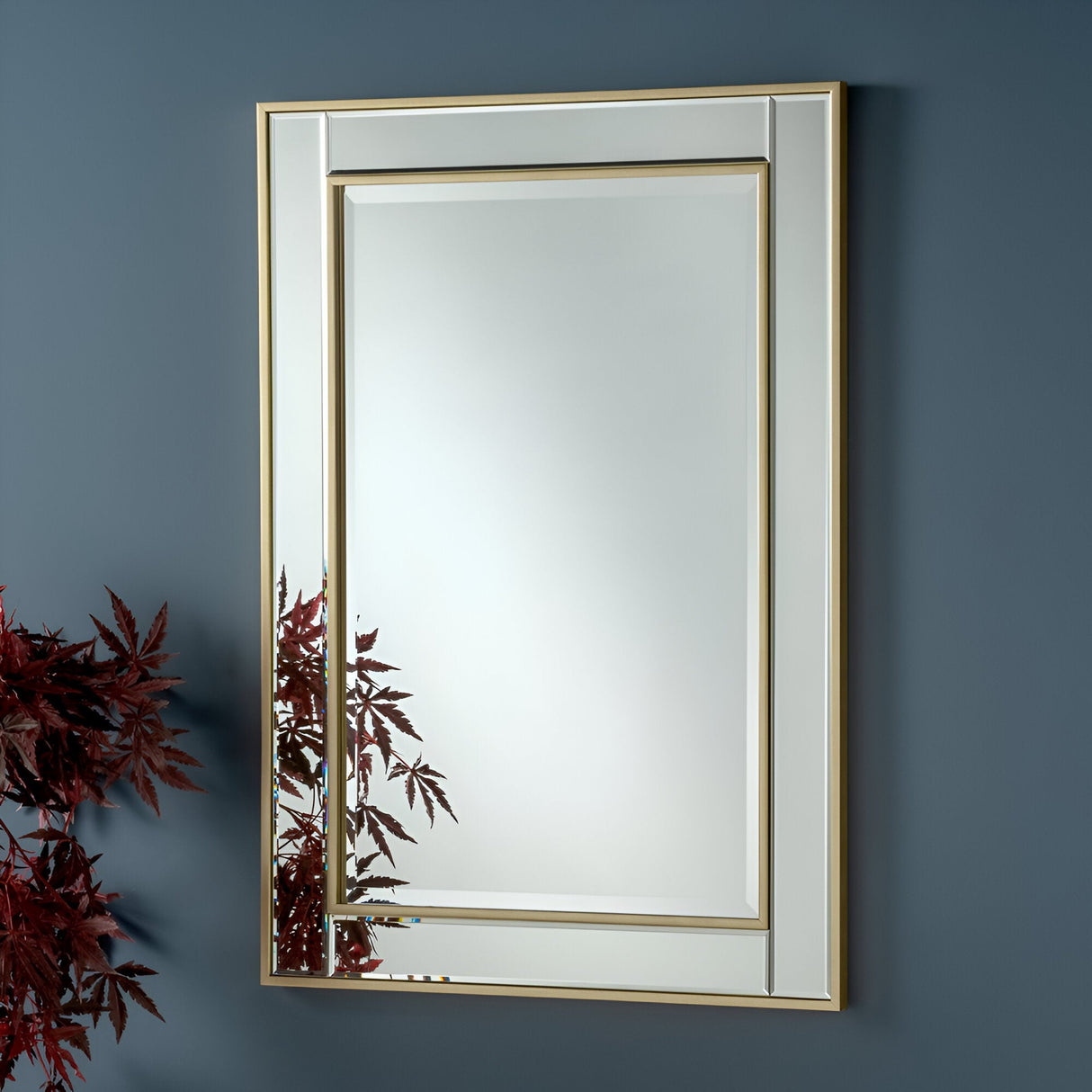 The Roma Rectangle Wall Mirror - Gold features an elegant double gold frame and is beautifully displayed against a dark teal wall. Red leaves from a surrounding plant gently encroach into the lower-left corner, casting their reflection in this luxurious decor piece.