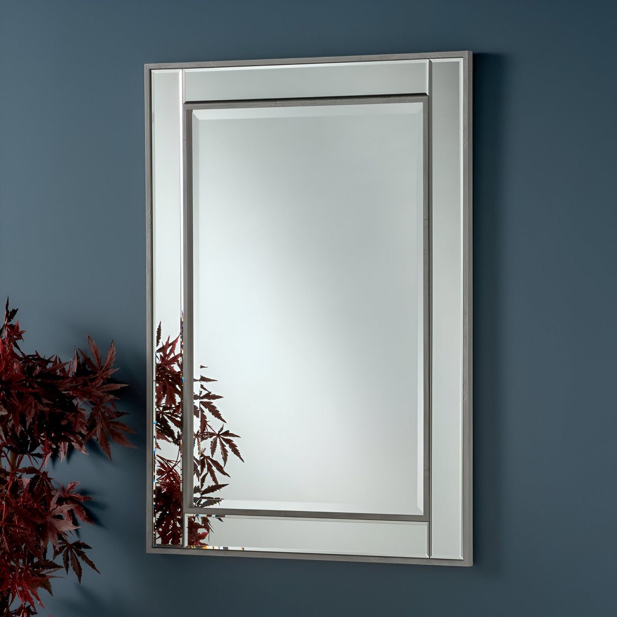 The Roma Rectangle Wall Mirror - Grey, a stunning piece of contemporary decor, features a double silver frame and is stylishly mounted on a dark blue wall. In the bottom left corner, the mirror beautifully reflects vibrant red maple leaves.