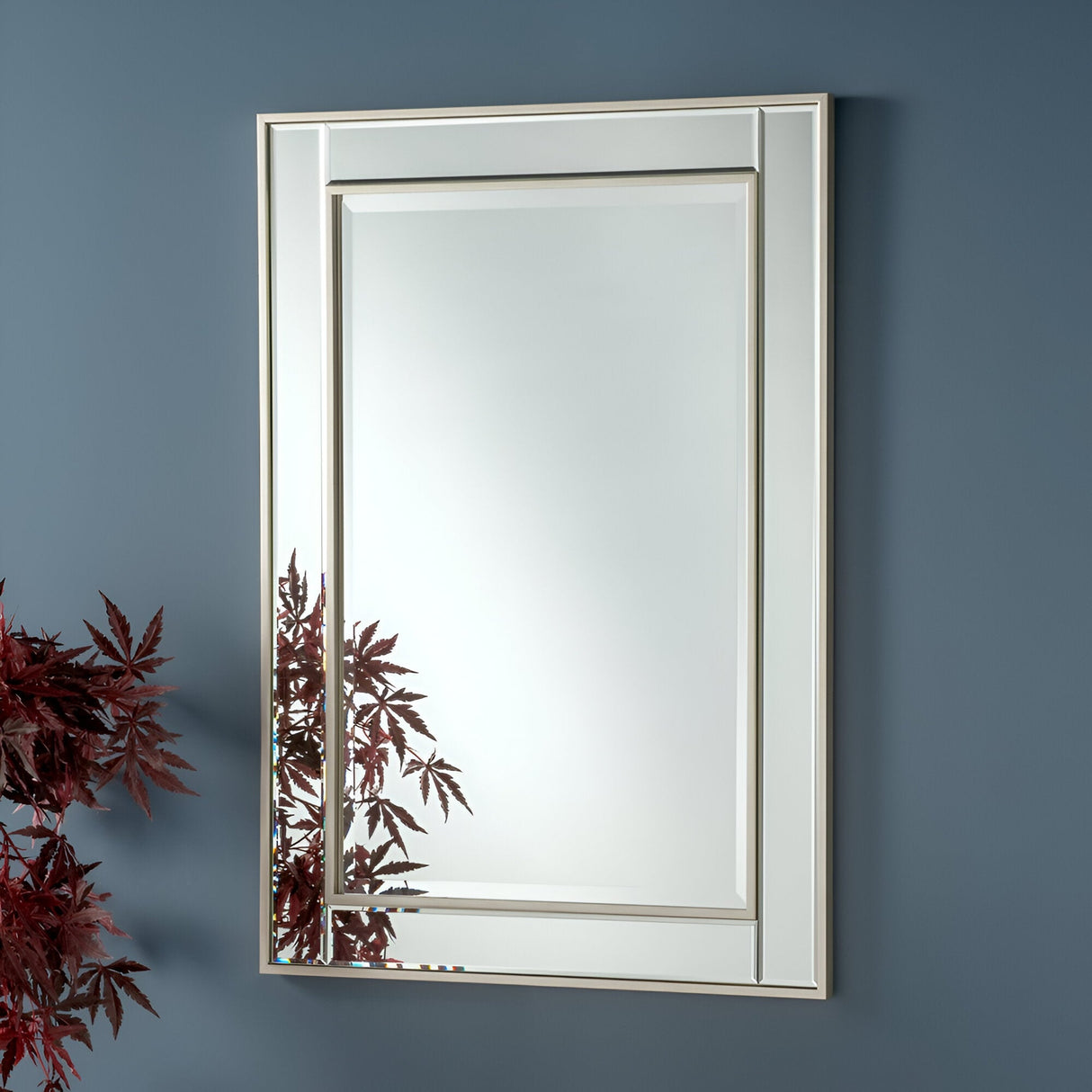 The Roma Rectangle Wall Mirror - Silver, featuring a beveled edge and silver frame, elegantly mirrors the vibrant red maple leaves against a blue wall. The partially visible leaves in the bottom left corner of the mirror add a natural element to the contemporary decor of this minimalist interior.