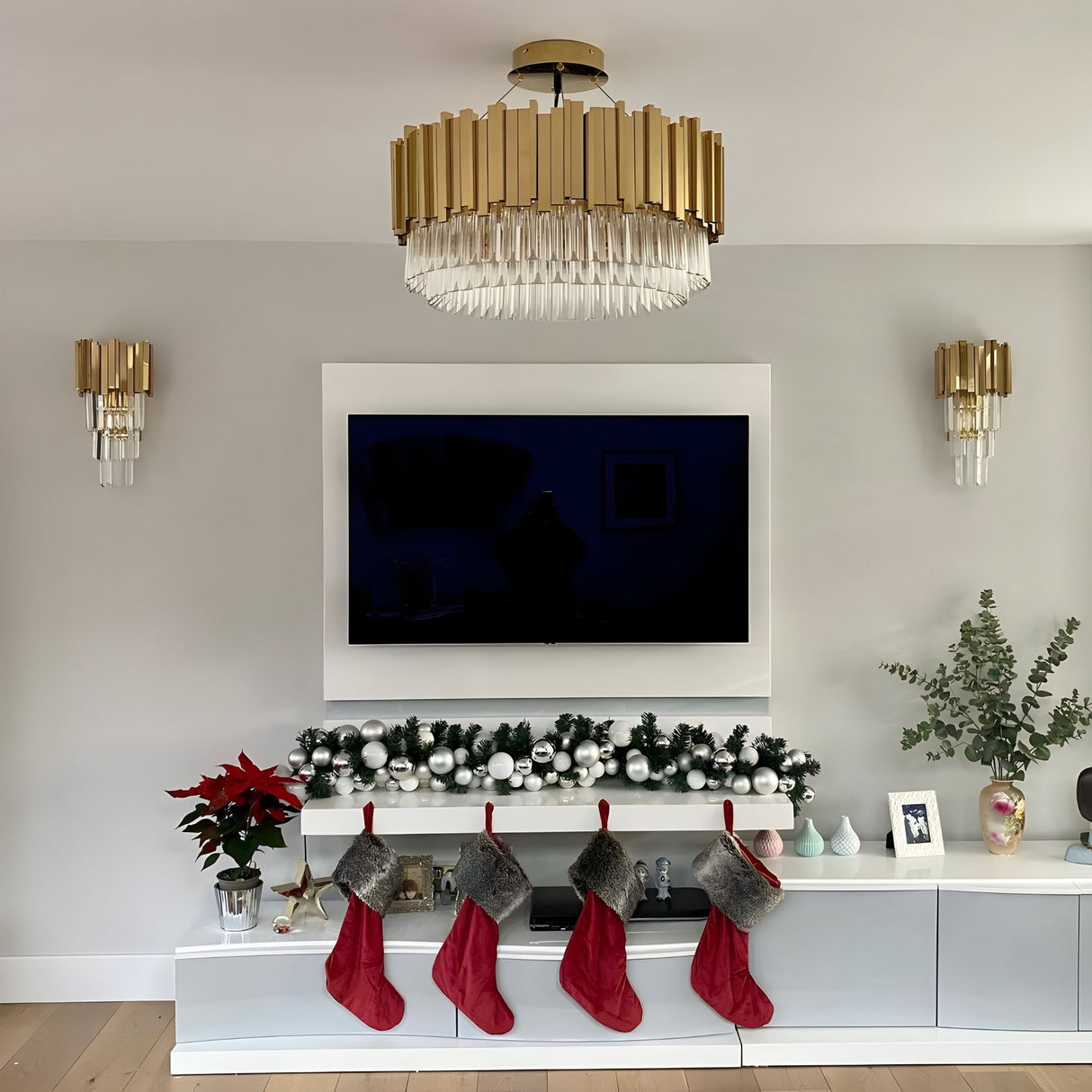 A modern living room with a TV on a grey wall, a Royal 6-12 Crystal Light Chandelier - Gold, four red Christmas stockings on a garland-decorated shelf, and festive ornaments, plants, and framed photos on the cabinet below.