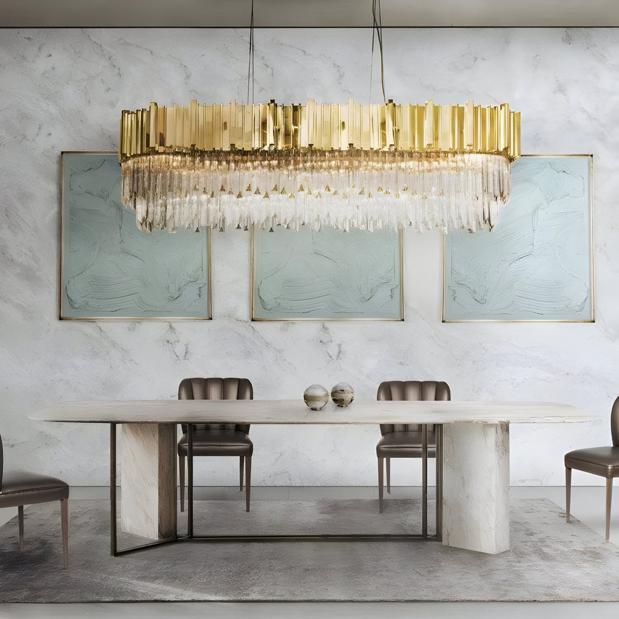 A modern dining room features a marble table, four brown chairs, three abstract paintings on a white marble wall, and the Royal 8-16 Light Crystal Chandelier - Gold. Two decorative spheres on the table enhance the luxurious interior.