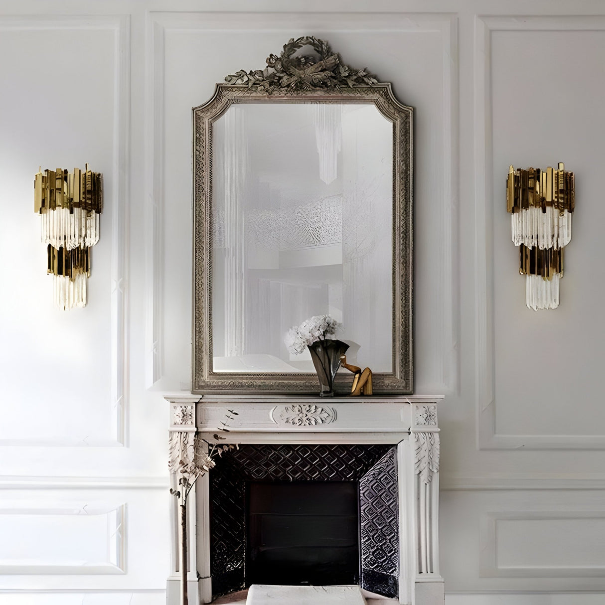 Elegant white fireplace with ornate detailing, topped by a large mirror. On the mantel sit a black vase with white flowers and a gold figurine. Royal Crystal Wall Light - Gold fixtures with crystal accents flank the mirror.