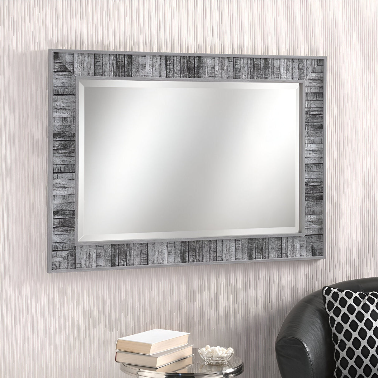 The Mirina Rectangle Wall Mirror in grey hangs on a white textured wall. Below it, a small table displays a stack of two books and a white bowl. On the right, you can partially see a black leather sofa adorned with a geometric patterned cushion.