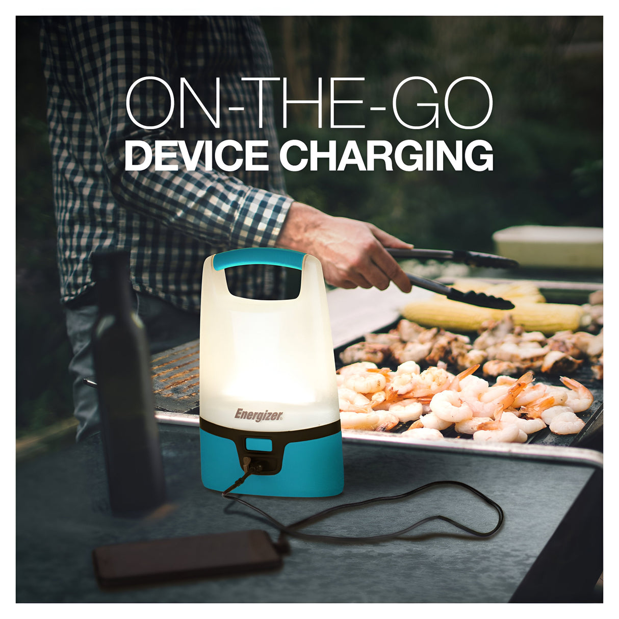 On an outdoor table, the Energizer Vision Rechargeable Lantern 1250 Lumen casts its bright light while charging a smartphone, setting the scene for a barbecue where someone grills shrimp and corn in the background.