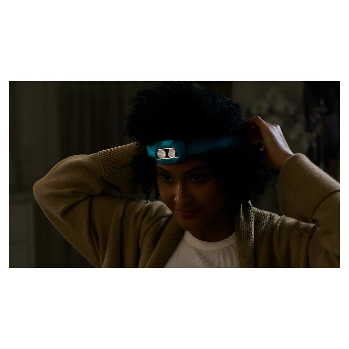 A person with curly hair is adjusting their Energizer LED Head Torch 250 Lumen - Batteries Included, preparing for hands-free illumination. Indoors in dim lighting, they are dressed in a light-colored sweater over a white shirt, ensuring that the headlamp fits securely on their head.