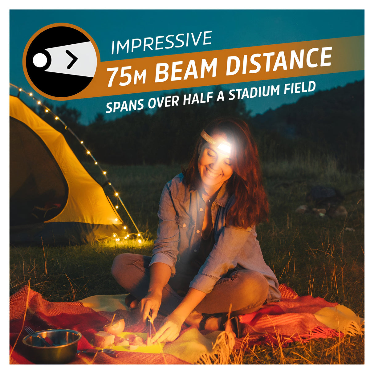 A person sits on a blanket near a tent, wearing an Energizer Waterproof LED Head Torch 500 Lumen in Orange. This powerful headlamp features an impressive 75-meter beam distance that spans over half a stadium field, making it ideal for outdoor camping at dusk. Additionally, its IPX7 waterproof rating ensures durability in any weather conditions.