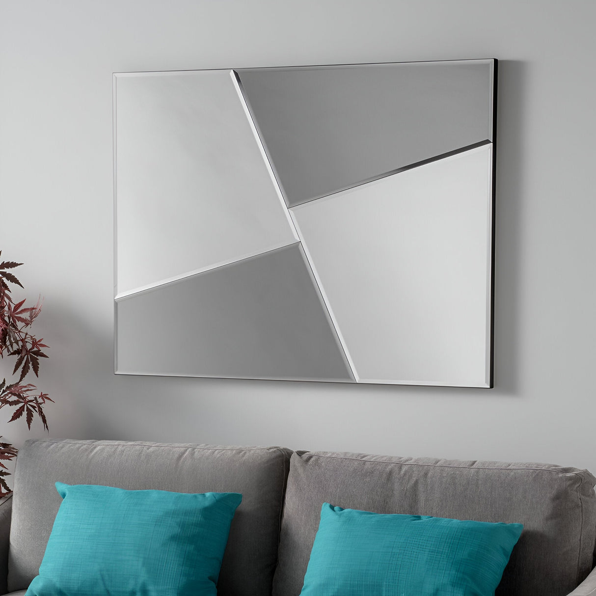 The Glaiza Rectangle Wall Mirror in Silver/Grey, measuring 122x91cm, adds a chic touch to the modern design of a minimalist space. It hangs on a light gray wall above a gray couch decorated with turquoise pillows, with a potted plant featuring red leaves partially visible on the left.
