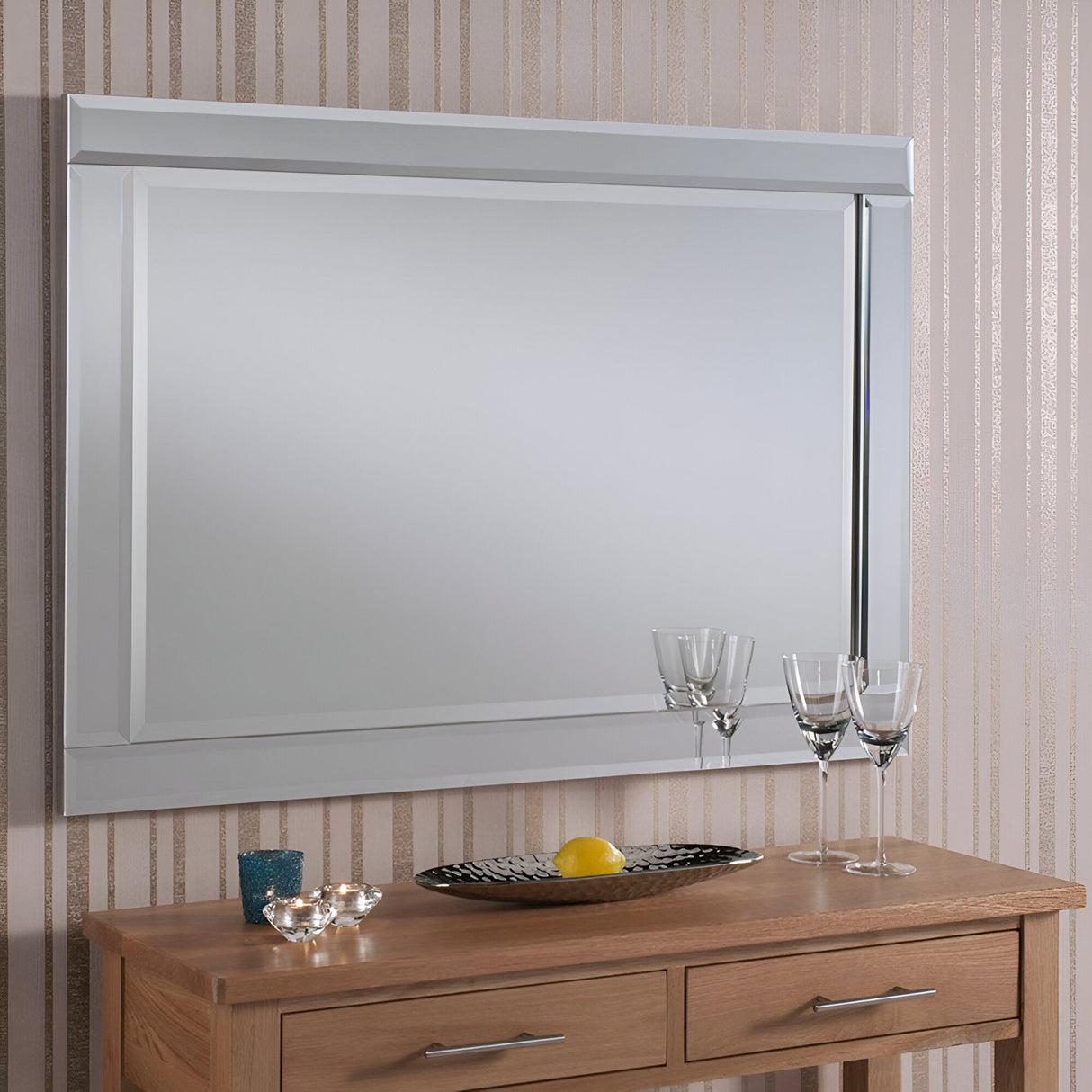 A Lustis Rectangle Wall Mirror - 104x74cm with a beveled edge is mounted on a wall adorned with beige-striped wallpaper, seamlessly enhancing the home's decor. Below it, a wooden console table holds three champagne glasses, a decorative candle, and a small fruit bowl.