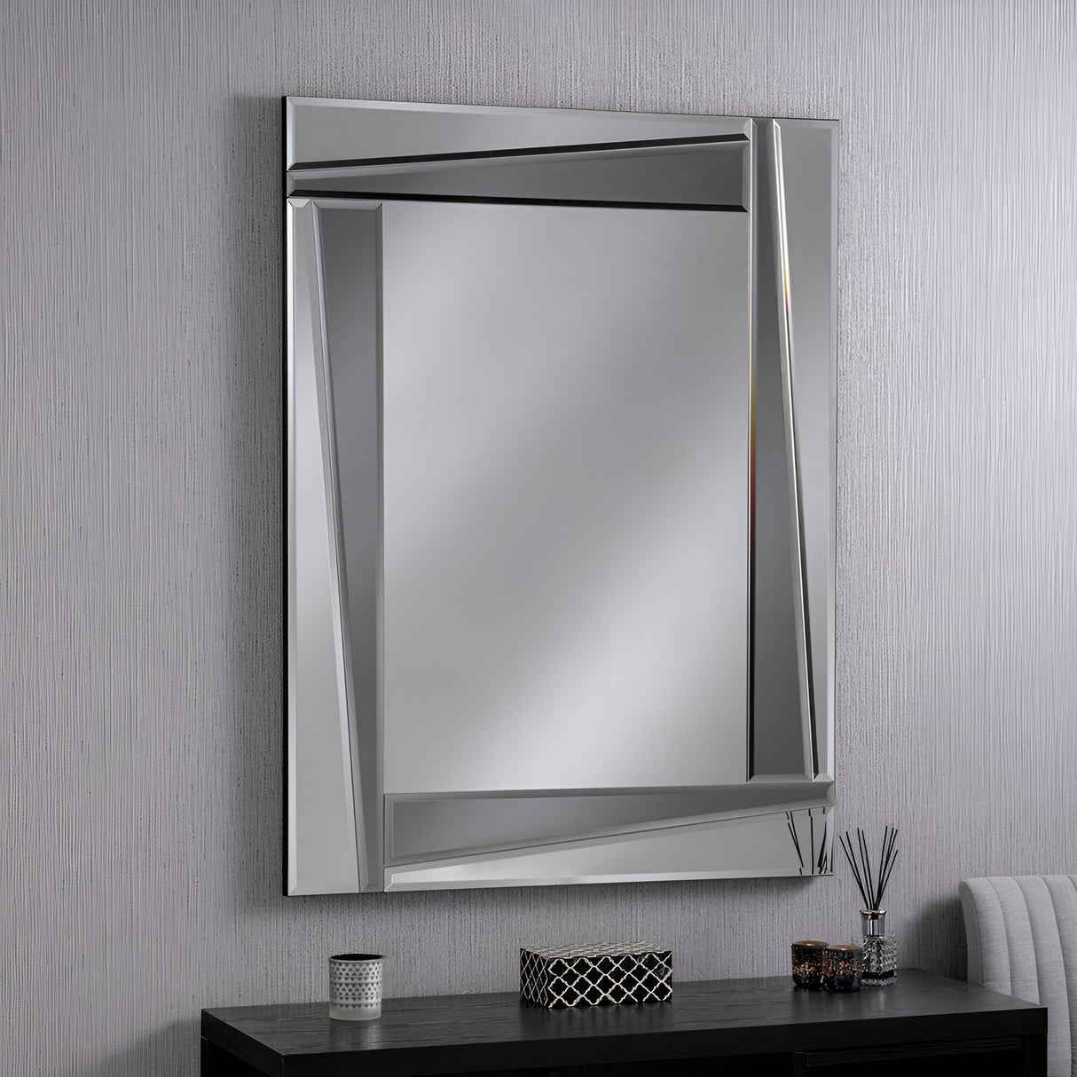 The Elara Rectangle Wall Mirror in Silver/Grey (122x92cm), featuring its angular, geometric frame, brings a hint of modern elegance to the space above a black table holding a decorative candle holder, sleek reed diffusers, and a small pot. The wall is subtly textured, making it ideal for contemporary interiors.