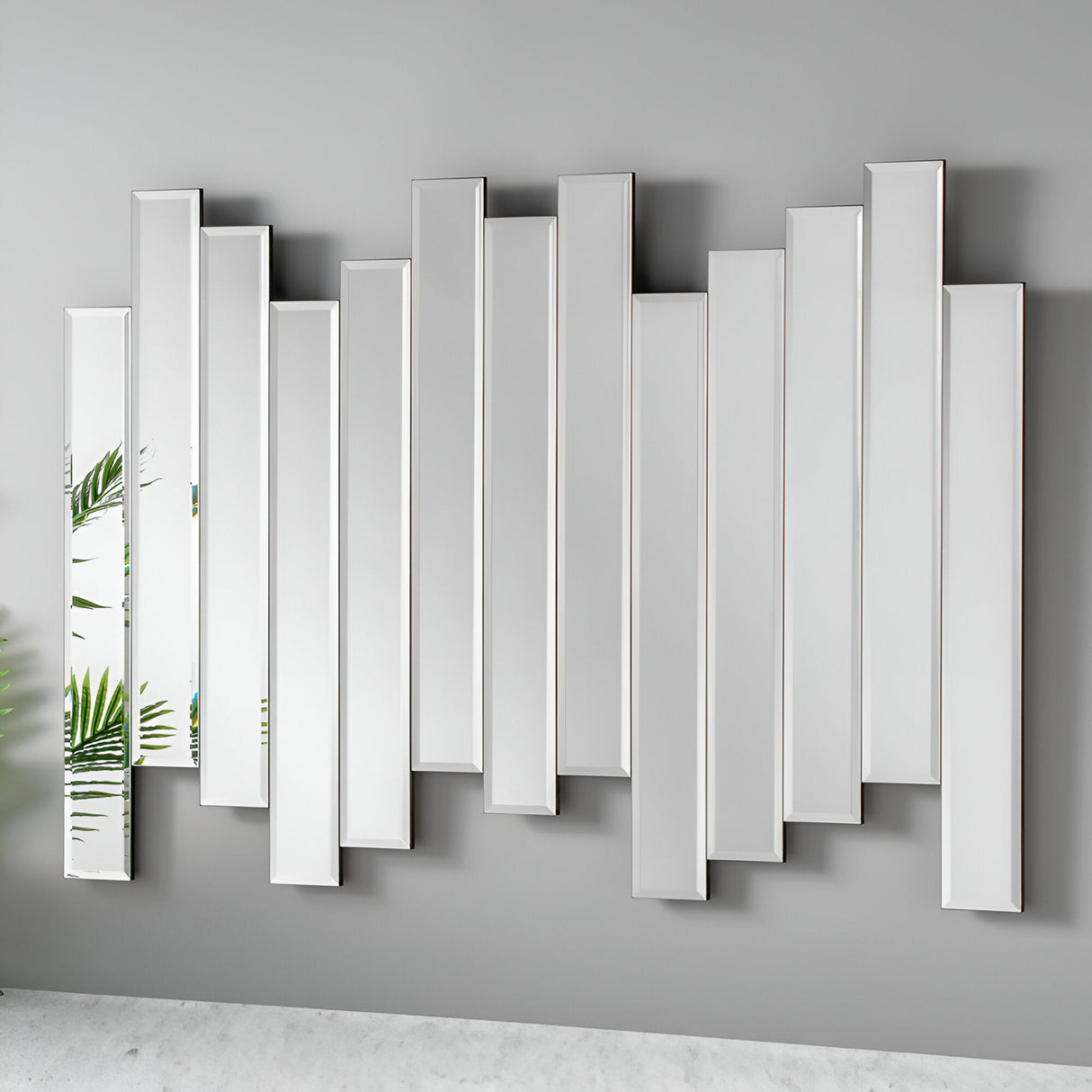 The Levira Panel Wall Mirror - Silver showcases a collection of staggered rectangular mirrors in a contemporary design with an elegant silver finish. Set abstractly against a light gray wall, it gorgeously captures and reflects the subtle hues of green from nearby plants, bringing a touch of natural color into the space.