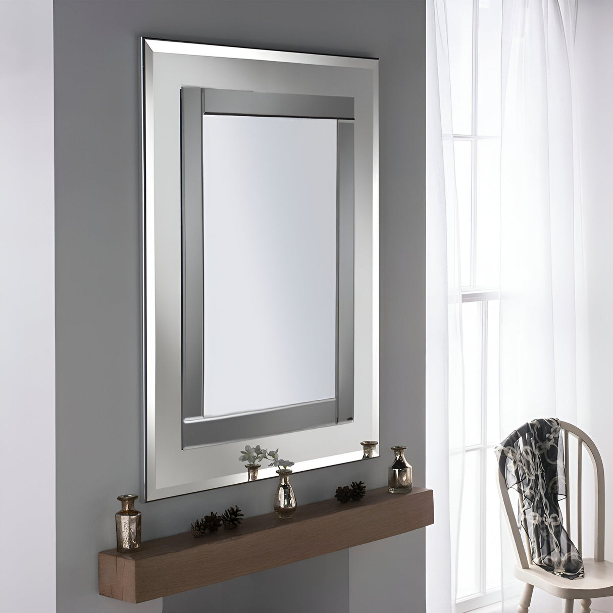 A large, modern luxury Salerno Rectangle Wall Mirror Grey/Silver- 103x77cm is mounted on a gray wall. Below it, a wooden mantel showcases sleek decorative items like silver candle holders and a small plant. Nearby, a white wooden chair with draped fabric complements the sheer curtains adorning the window.