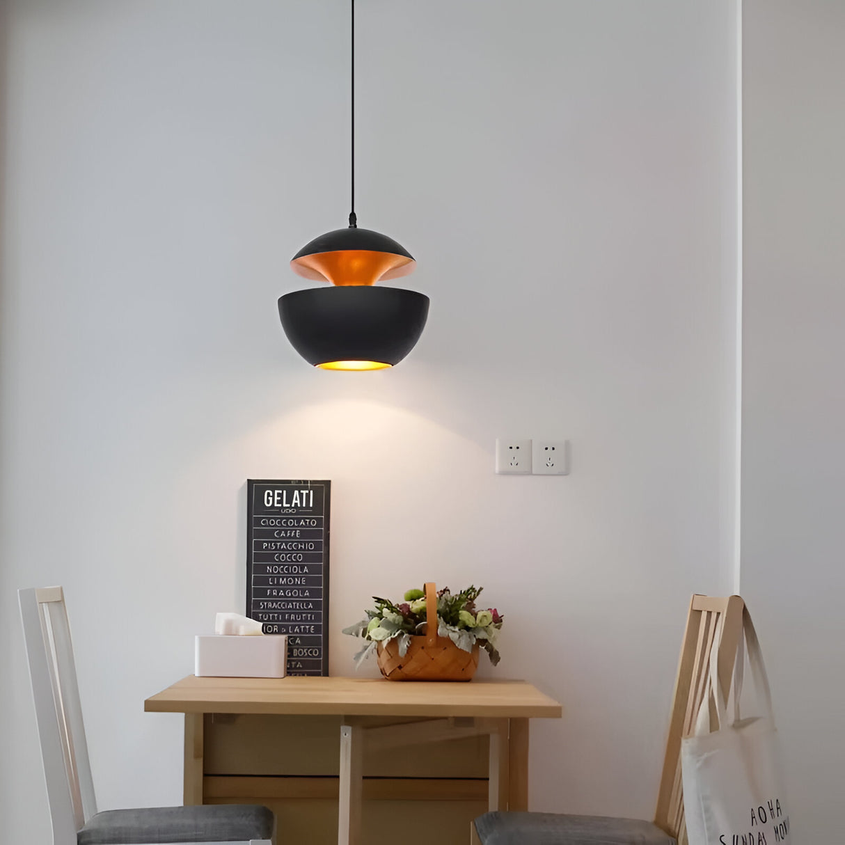 A minimalist dining area features a wooden table with two chairs, alongside a decorative flower basket and a menu board listing Gelati. Overhead, an Ex-Display Core Pendant Copper & Black - 35cm illuminates the scene. A bag hangs from one chair, completing the cozy space.