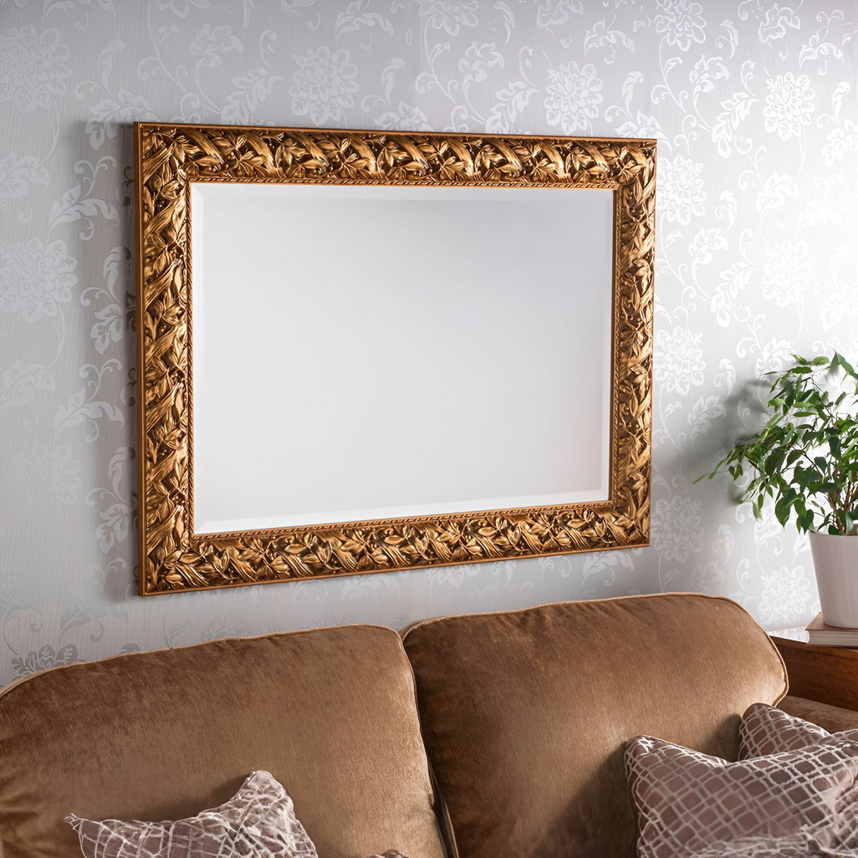 A large, ornate Evixa Rectangle Wall Mirror in dark gold is mounted on a patterned wallpapered wall above a brown sofa adorned with patterned cushions. A green plant sits on a wooden side table beside the sofa, enhancing the luxurious home decor.