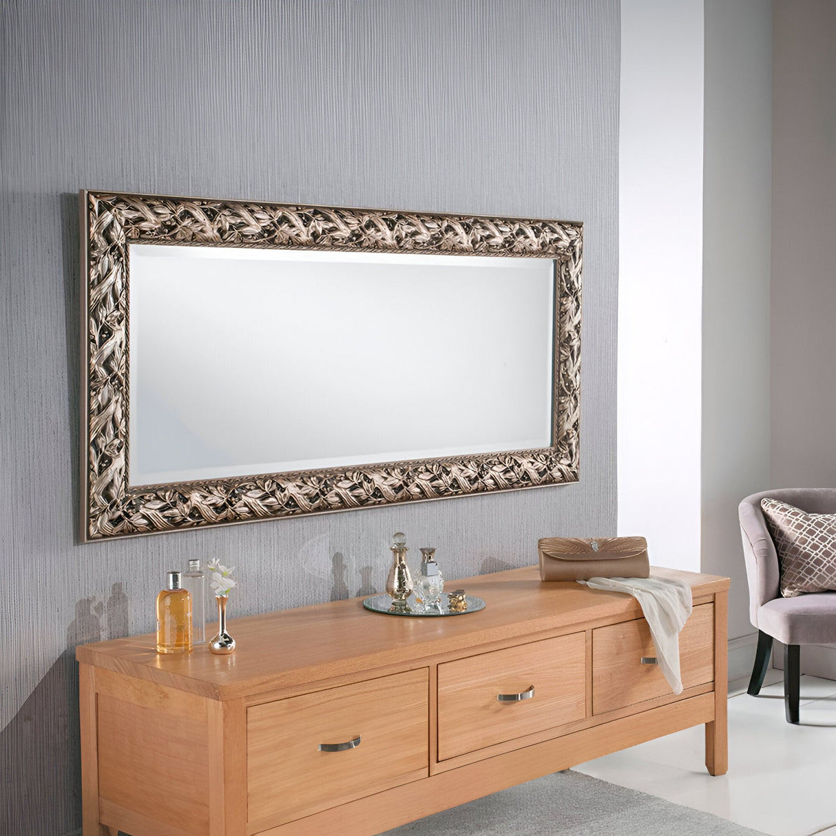 An Evixa Rectangle Wall Mirror - Silver is mounted above a wooden console table with drawers. The tabletop features decorative items, such as a tray with bottles and a small vase. To the right, a gray cushioned chair partially comes into view, enhancing the luxurious home decor setting.