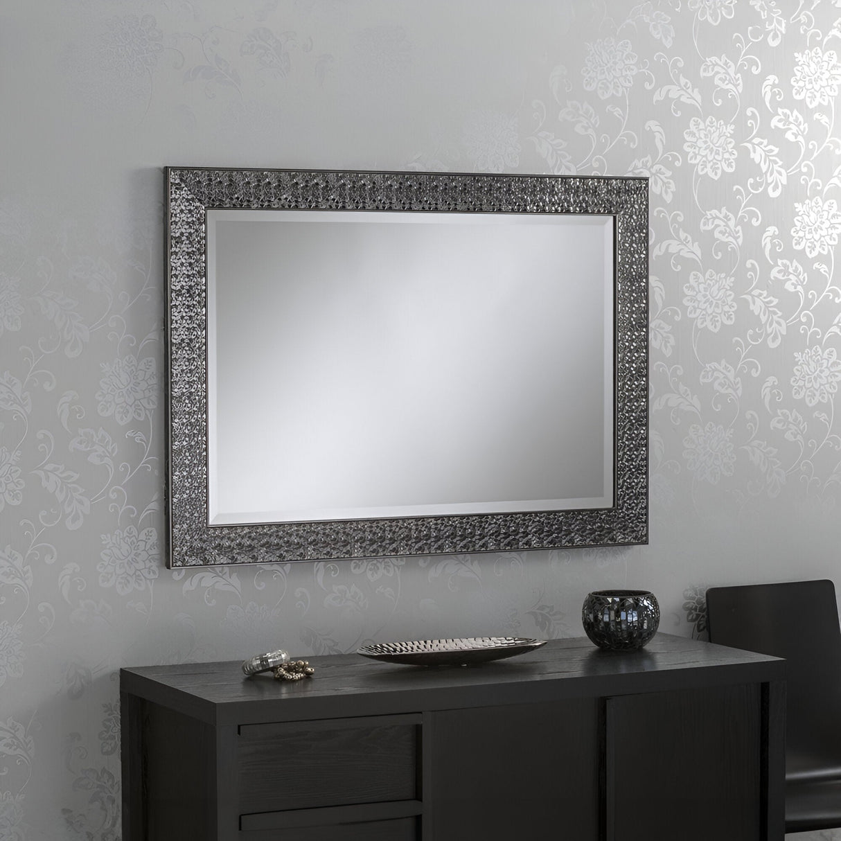 A Siena Rectangle Wall Mirror in grey, featuring an ornate, textured frame, hangs on a patterned light gray wall, adding a contemporary touch. Below it sits a dark wood console table adorned with a decorative bowl and various small items. Part of a black chair is visible on the right.