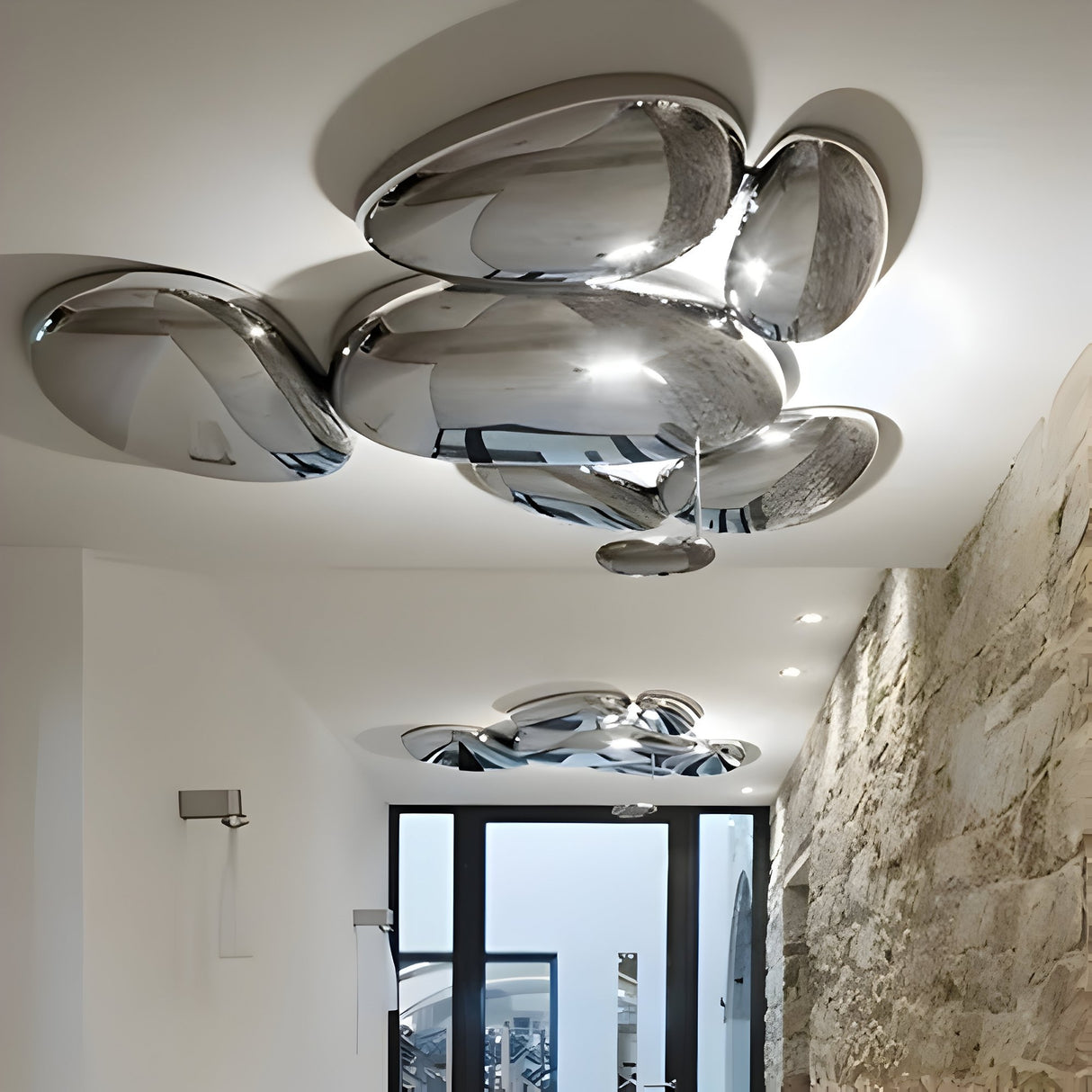 Modern interior featuring abstract, shiny Skydro 5/6 LED Ceiling Light - Silver fixtures on a white ceiling. Below, a stone wall lines one side, with large glass doors at the hallways end letting in natural light.