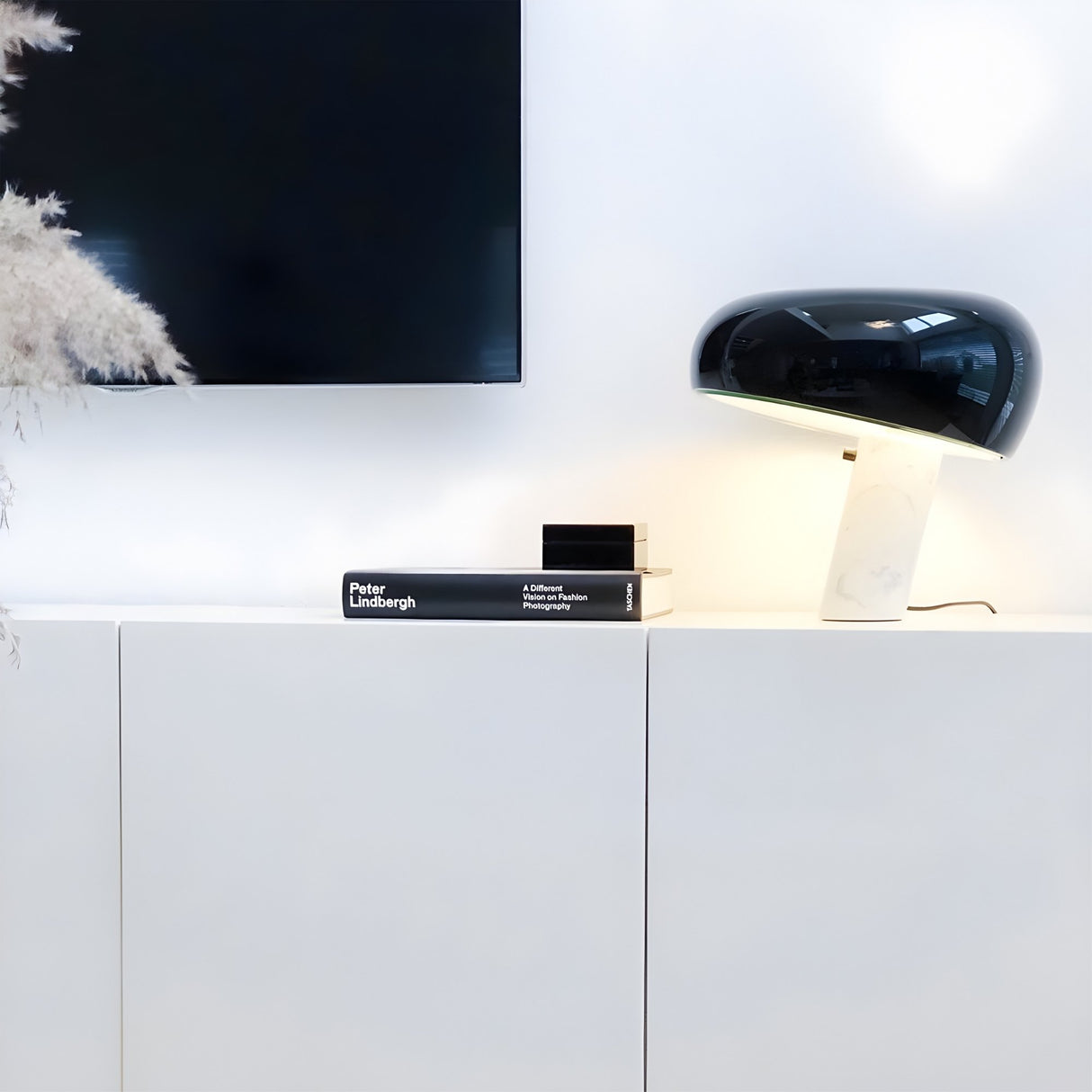 Niori Sleepy Marble Table Lamp with black glass dome shade and white marble base on a white sideboard beside a Peter Lindbergh book, below a wall-mounted TV.