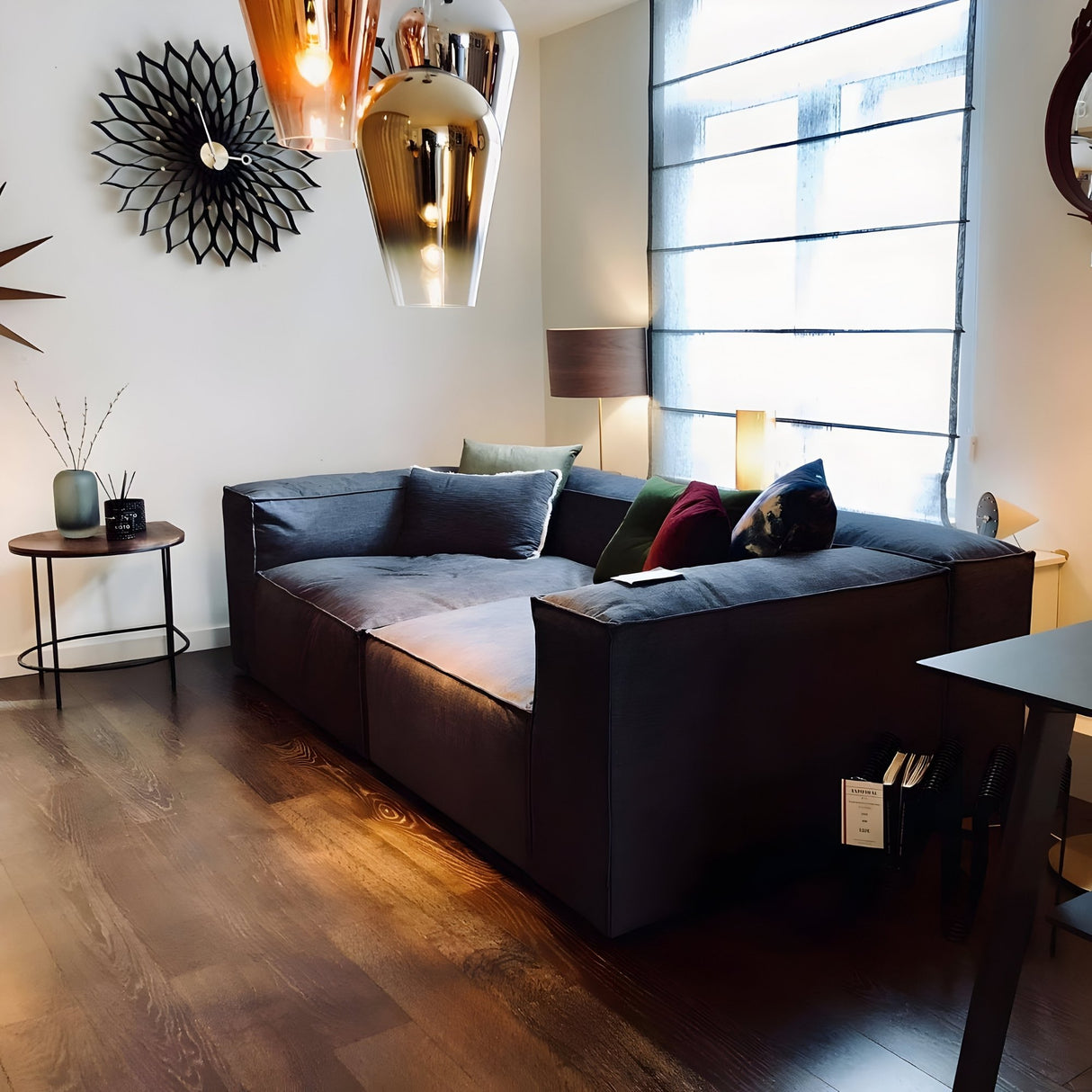 A modern living room with a dark grey sofa, colorful pillows, a round side table, wooden floors, large wall mirrors by a window with sheer blinds, and the Slice Glass Pendant Light - Gold as handcrafted lighting.