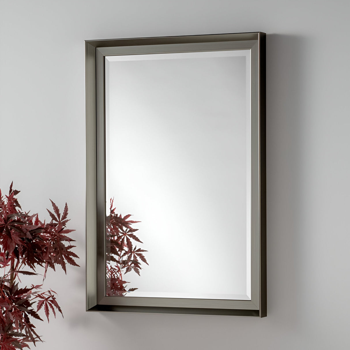 The Sloane Rectangle Wall Mirror - Grey is mounted on the wall, reflecting a red-leafed plant in the bottom left corner and enhancing the room's bright and minimalistic appeal. Its modern decor effortlessly complements any space.