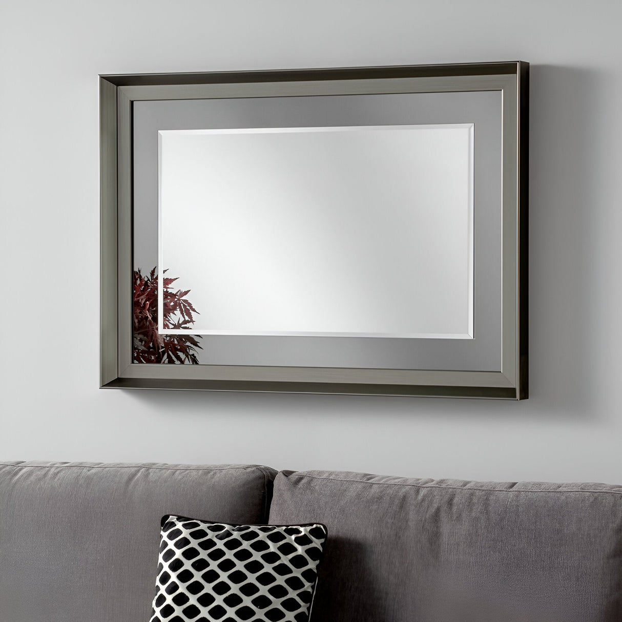 A Sloane Rectangle Wall Mirror with a sleek, modern sophistication is mounted on a light gray wall. Below the mirror, a gray sofa adorned with a black and white patterned cushion adds charm. The reflection of red leaves in the mirror brings subtle vibrancy to the serene space.