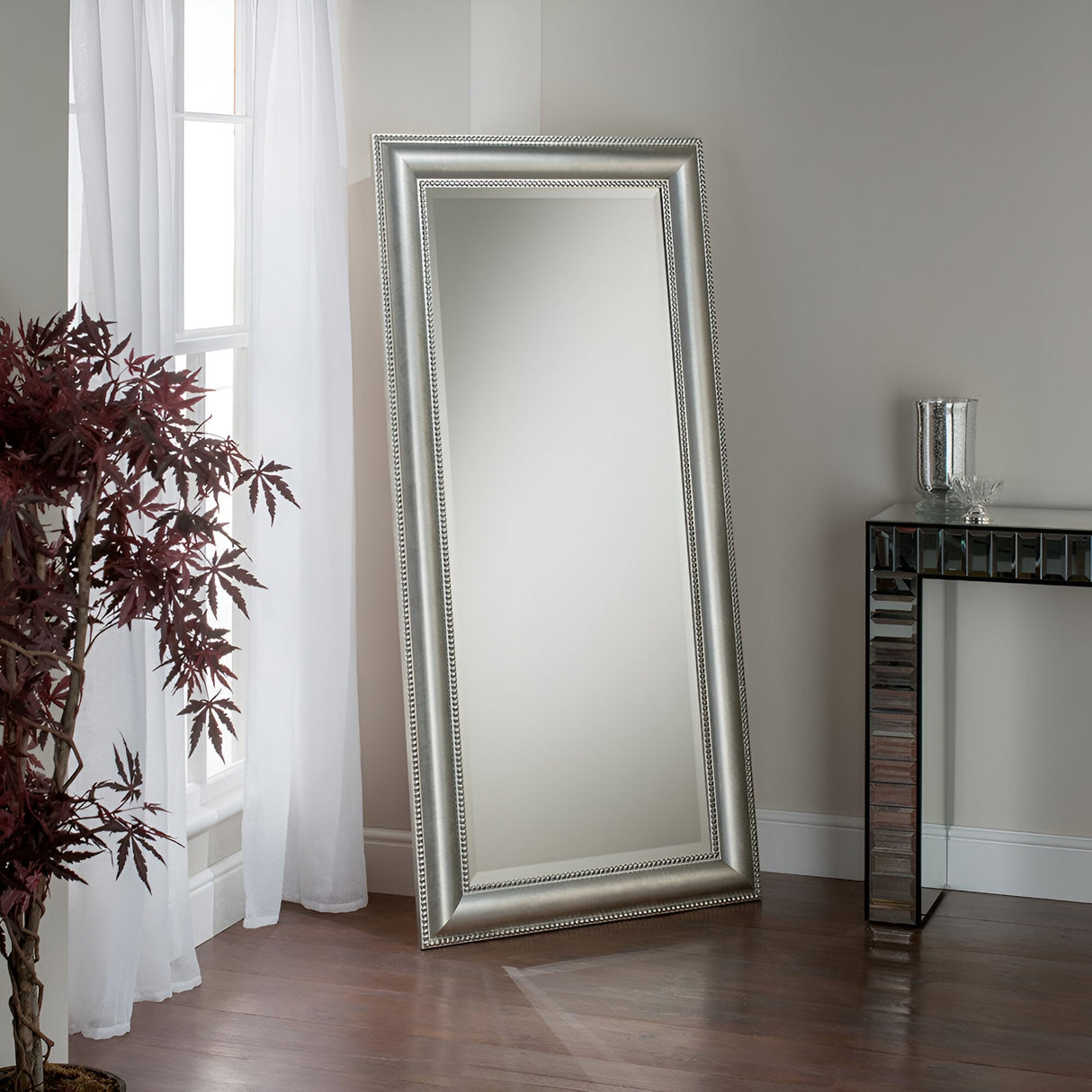 The Sorrento Leaner Wall Mirror - Silver is placed on the floor against a light-colored wall. A decorative plant and a glass-topped table complement the contemporary decor on the wooden floor, while sheer curtains over a window let natural light flood into the room.