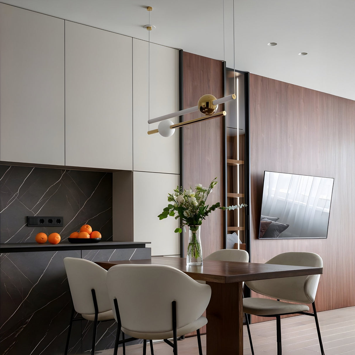 Modern kitchen and dining area with a wooden table, cream chairs, flowers in a vase, oranges on a dark marble countertop, minimalist cabinets, wood-paneled wall, and wall-mounted TV. The Sottile LED Horizontal Suspension Light - Gold enhances the sleek design above the table.