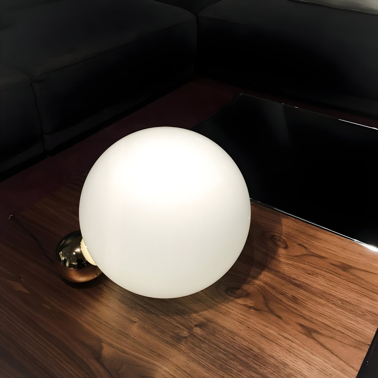 The Soul Table Lamp - Copper, featuring a large white frosted glass globe and a copper-finish base, is placed on a wooden table beside a black sofa and a glossy black surface.